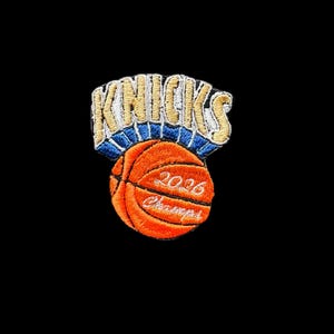 May include: An embroidered patch featuring the word KNICKS in yellow with a blue outline, positioned above an orange basketball. The basketball includes the white embroidered text 2026 and the word Champions in a cursive script font.