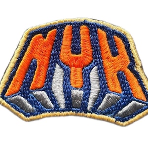 May include: An embroidered patch featuring the letters NYK in bright orange thread against a dark blue background. Below the letters are silver-colored vertical shapes, and the entire patch is outlined with a thick, light tan border.