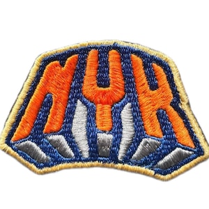 Puede incluir: Un parche bordado que presenta las letras NYK en hilo naranja brillante sobre un fondo azul oscuro. Debajo de las letras hay rayos verticales de color gris plateado, y toda la forma está delineada con un borde grueso de color amarillo claro.