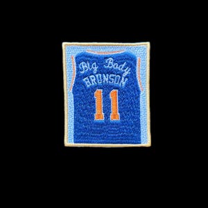 Op de afbeelding: Een rechthoekige geborduurde patch met een blauw basketbalshirt-ontwerp met oranje accenten. De tekst Big Body BRUNSON is in het wit geborduurd boven het oranje nummer 11. De patch heeft een lichtblauwe rand en een crèmekleurige buitenrand.