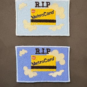 May include: Two embroidered patches with a light blue background and cream-colored clouds. The patches feature a yellow MetroCard with the text "R.I.P" above it. The MetroCard has black stripes and the words "MetroCard" in blue.