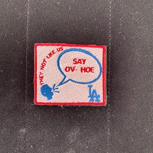 Op de afbeelding: Een geborduurde patch in rood en blauw met de tekst "They Not Like Us" en "Say Ove Hoe" in een spreekballon. De patch heeft ook een gestileerd LA-logo.
