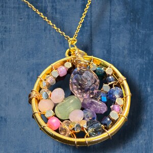 Puede incluir: Un collar de oro con un colgante redondo lleno de varias piedras preciosas de colores, incluyendo rosa, morado, verde y azul.