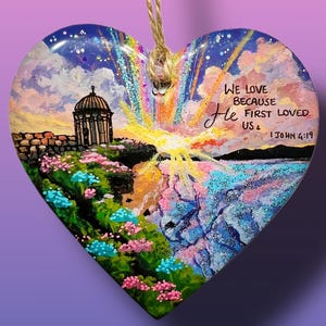 May include: A heart-shaped ornament with a painted scene of a sunset over a body of water. The ornament features a rainbow, a building with a dome, and flowers. The text "We love because He first loved us 1 John 4:19" is written on the ornament.