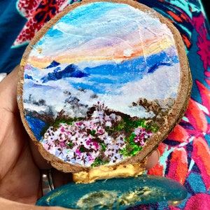 Peut inclure: Un disque en bois peint à la main représentant un paysage de montagnes, de nuages et de fleurs. Le disque est monté sur une base bleue avec des accents dorés.