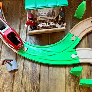 May include: A wooden train set with a red train, green track, and a wooden train station with the word "BRIO" on it. There are also two wooden figures, one wearing a red hat and the other wearing a brown dress.