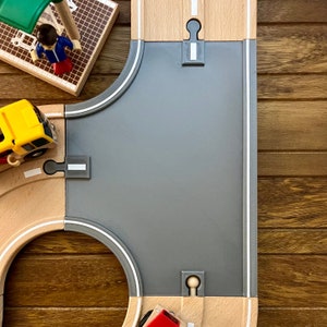 May include: A wooden toy road intersection with a gray asphalt surface. The intersection has a curved road on the left and a straight road on the right. There is a yellow toy bus on the curved road and a red toy car on the straight road.