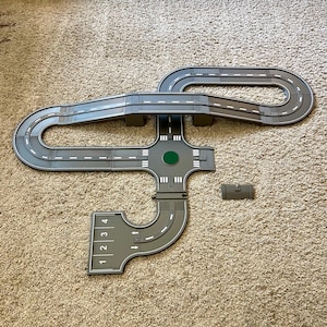 Puede incluir: Un sistema de carreteras de juguete de plástico gris con un puente, una intersección de cuatro vías y una sección curva con los números del 1 al 4 impresos en ella.
