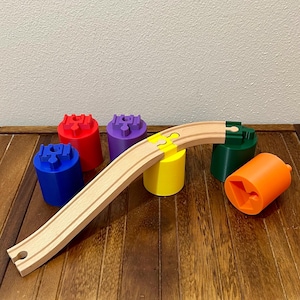 Könnte beinhalten: Eine hölzerne Spielzeug-Eisenbahnstrecke mit gebogenem Design, begleitet von farbenfrohen zylindrischen Stützen in Rot, Blau, Lila, Gelb, Grün und Orange. Die Strecke ist hellbraun, die Stützen haben einzigartige Designs.