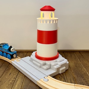 May include: A red and white lighthouse with a light on top, sitting on a grey rock base. A blue toy train is in the foreground.