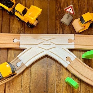May include: Wooden toy train set with a white intersection piece, wooden tracks, yellow construction vehicles, green trees, and a road sign. The set is arranged on a wooden surface.