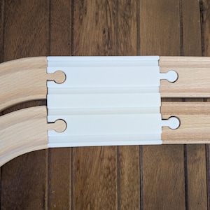 Puede incluir: Una vía de tren de madera blanca con dos vías curvas de madera a cada lado. La vía tiene un inserto de plástico blanco con tres ranuras.