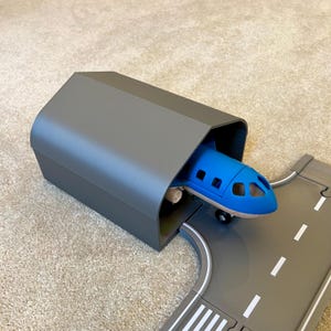 May include: A blue toy airplane emerges from a gray hangar, positioned on a gray runway with white markings. The toy airplane has a blue fuselage and white wheels. The hangar is a simple, rectangular structure.