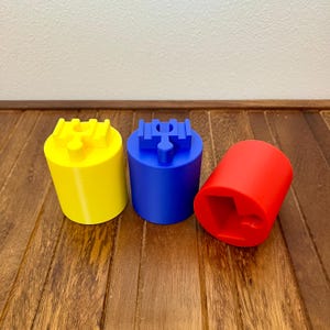 May include: Three brightly colored, cylindrical 3D-printed objects. A yellow cylinder with a cross-shaped top, a blue cylinder with a similar top, and a red cylinder with a square-shaped opening. The objects are on a wooden surface.
