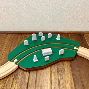 Puede incluir: Una escena de tren de juguete de madera con una isla verde, pequeñas lápidas blancas y un ataúd. La vía es de madera clara, colocada sobre una superficie de madera oscura, evocando un tema de cementerio.