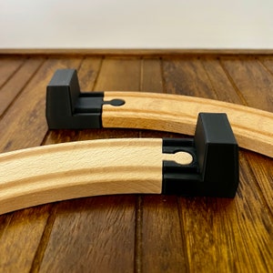 Puede incluir: Dos piezas de vía de tren de madera con conectores de plástico negro. Las piezas de vía son curvas y de madera clara.