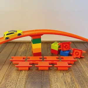 May include: A yellow toy car on an orange track, with a colorful block tower supporting the track. Additional orange track pieces and colorful blocks are scattered on a wooden surface. A fun toy set for kids.