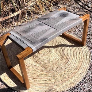 Può includere: Panca in legno con seduta grigia intrecciata. La panca ha una forma rettangolare con struttura in legno naturale ed è posizionata su un tappeto rotondo intrecciato. La panca è lunga circa 122 cm.