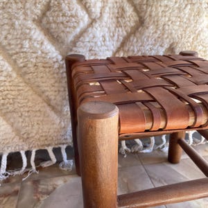 Puede incluir: Un taburete de madera artesanal con un asiento de cuero tejido. El taburete presenta cuatro patas de madera robustas y un acabado marrón cálido. El asiento está hecho de tiras de cuero entrelazadas, creando una superficie texturizada.