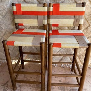 Puede incluir: Dos taburetes de bar de madera con asientos tejidos en naranja, beige y crema. Los taburetes tienen un diseño sencillo con cuatro patas y un solo peldaño.