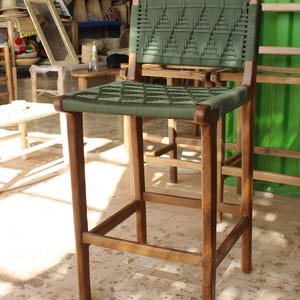 May include: A high wooden bar stool featuring a woven green seat and back. The frame is constructed from light brown wood. The stool has four legs and a footrest. The woven seat and back have a geometric design.