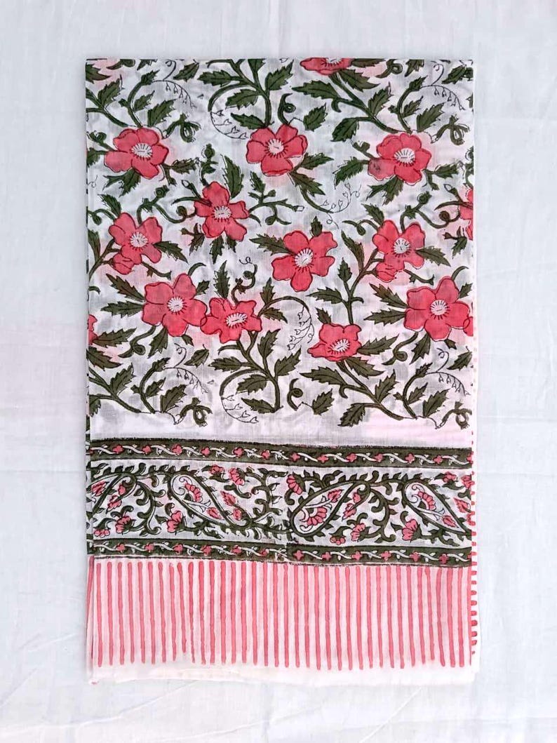 May include: White textile with a floral design of pink flowers and green leaves. The fabric has a border with paisley and pink vertical stripes. The textile is folded and displayed on a white surface.