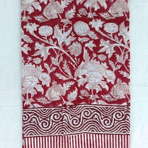 May include: A folded textile featuring a repeating floral pattern in white on a deep red background. The design includes large stylized flowers and foliage. The bottom border has a striped pattern and a wavy design.
