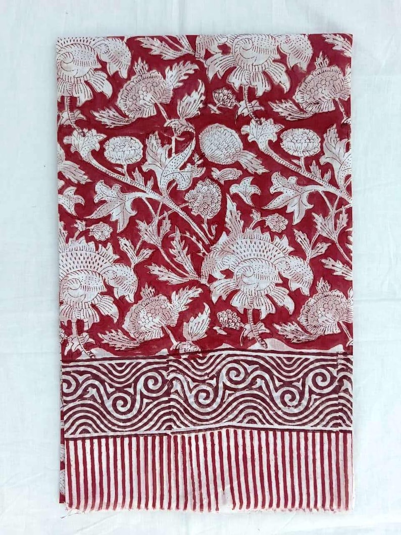 May include: A folded textile featuring a repeating floral pattern in white on a deep red background. The design includes large stylized flowers and foliage. The bottom border has a striped pattern and a wavy design.