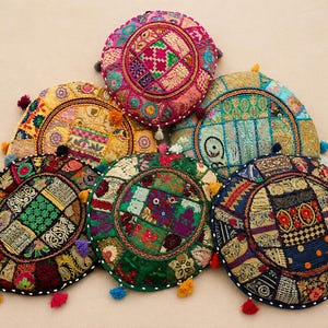 Könnte beinhalten: Sechs runde Bodenkissen mit komplexen Patchwork-Designs, bestickten Mustern, metallischen Garnakzenten und kleinen bunten Quasten an den Rändern. Die Kissen zeigen eine Vielzahl lebendiger Farben, darunter Pink, Gelb, Türkis, Grün und Dunkelblau.
