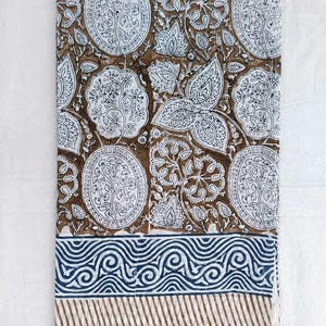 May include: A rectangular textile with a brown and white floral pattern and a blue and white border. The design features repeating floral motifs in white on a brown backdrop. The border has a blue wave pattern and brown stripes.
