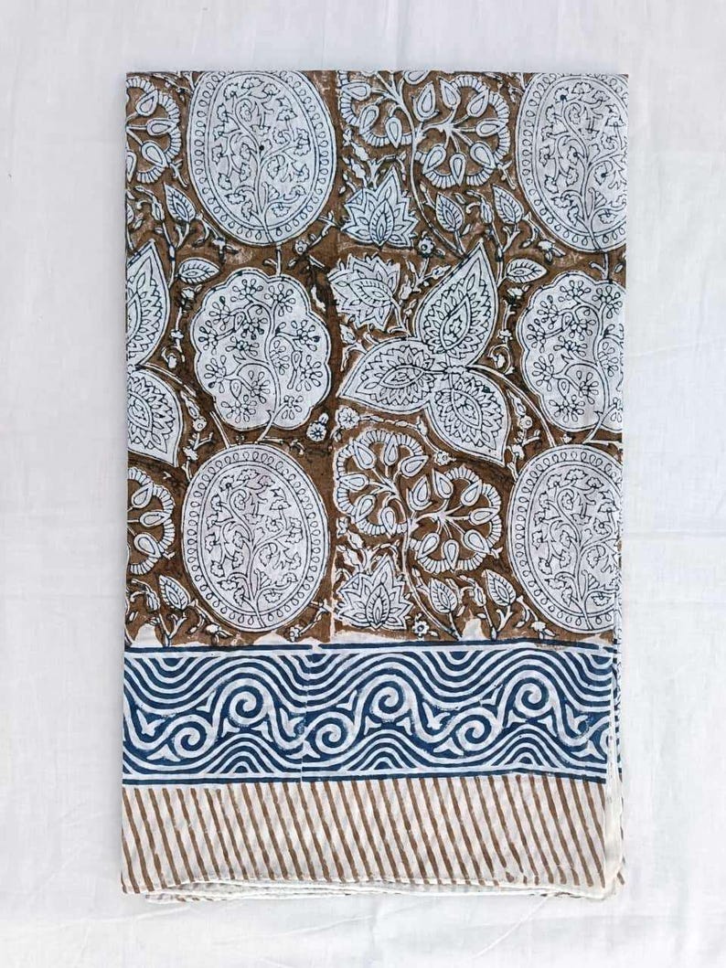 May include: A rectangular textile with a brown and white floral pattern and a blue and white border. The design features repeating floral motifs in white on a brown backdrop. The border has a blue wave pattern and brown stripes.