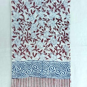 May include: A folded white textile with a floral pattern in blue and maroon. The design features stylized flowers and vines. The bottom border has a blue swirl pattern above maroon vertical stripes. The fabric is likely cotton or linen.