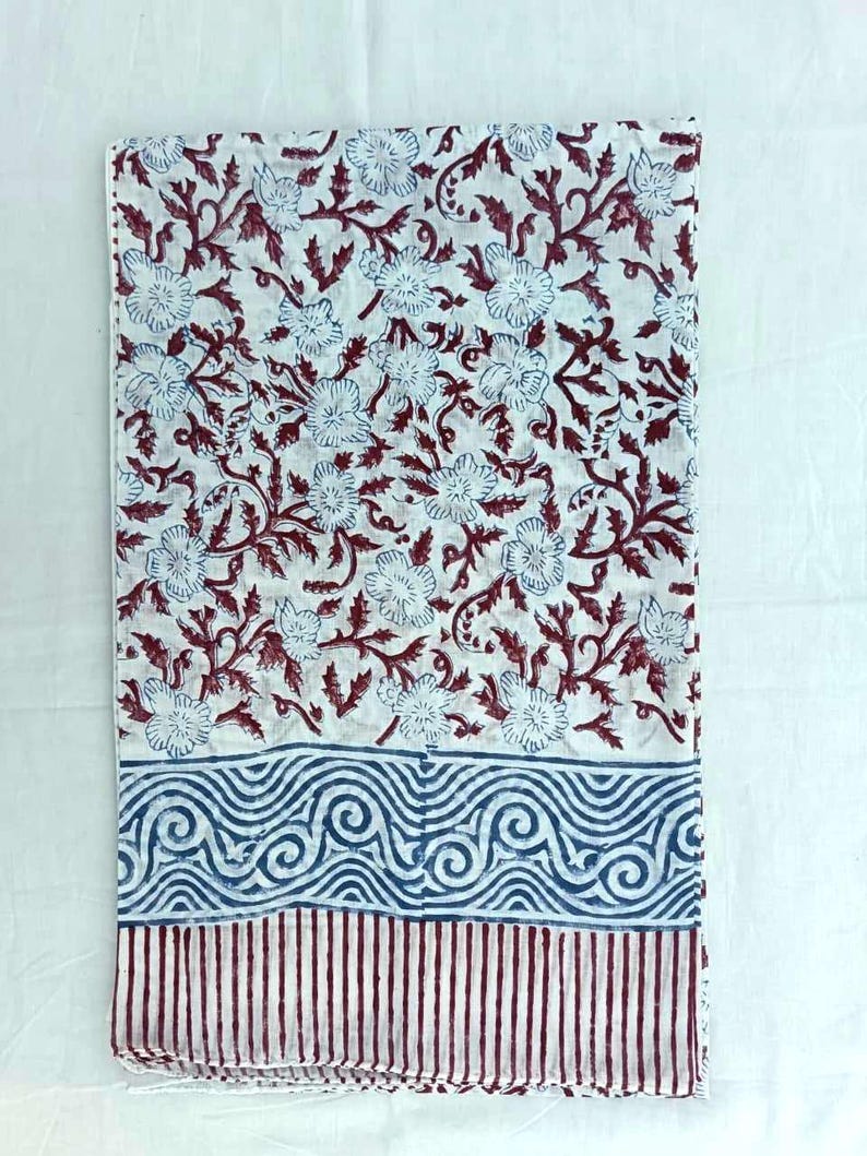 May include: A folded white textile with a floral pattern in blue and maroon. The design features stylized flowers and vines. The bottom border has a blue swirl pattern above maroon vertical stripes. The fabric is likely cotton or linen.