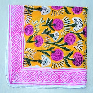 K&ouml;nnte beinhalten: Quadratischer, gefalteter Stoff mit leuchtend gelbem Hintergrund. Er zeigt ein sich wiederholendes Muster aus rosa und wei&szlig;en floralen Designs mit gr&uuml;nen Bl&auml;ttern. Ein rosa geometrischer Rand umrahmt die Kanten und schafft einen dekorativen Stoff.