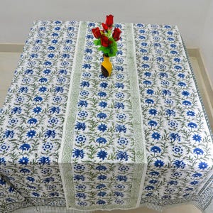 May include: A white tablecloth with a blue floral pattern and green accents. The tablecloth is rectangular and covers a table. A vase with red flowers sits on the table. The tablecloth has a border with a similar design.
