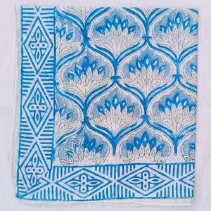 K&ouml;nnte beinhalten: Ein wei&szlig;es Textil mit einem sich wiederholenden blauen Blumenmuster und einem blauen geometrischen Rand. Das florale Design zeigt stilisierte Bl&uuml;ten in Blau- und Wei&szlig;t&ouml;nen. Der Rand enth&auml;lt Diamant- und Streifenmuster.