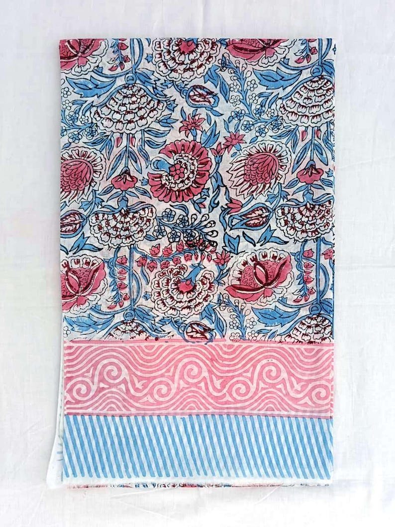 May include: A folded textile with a floral design in pink and blue on a white backdrop. The pattern showcases large flowers and leaves. The lower section has a pink wavy pattern and blue stripes.