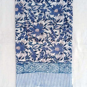 May include: A folded textile with a blue floral pattern on a white backdrop. The design incorporates large, stylised flowers, leaves, and vines. A border of geometric shapes and vertical stripes in blue accents the bottom edge. The fabric appears to be cotton or linen.