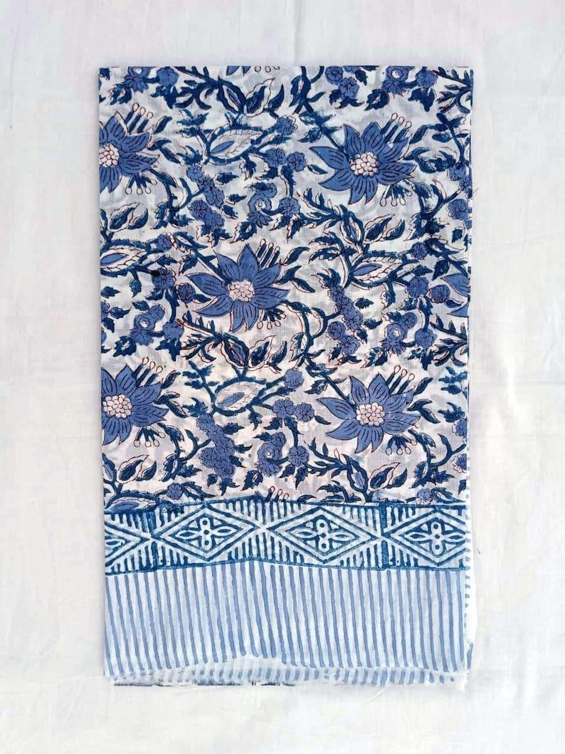 May include: A folded textile with a blue floral pattern on a white backdrop. The design incorporates large, stylised flowers, leaves, and vines. A border of geometric shapes and vertical stripes in blue accents the bottom edge. The fabric appears to be cotton or linen.