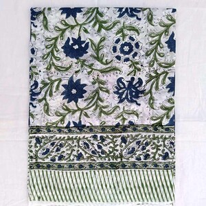 May include: A folded white textile with a repeating floral pattern in navy blue and green. The design features stylized flowers and leaves. A border with paisley and striped patterns is visible.
