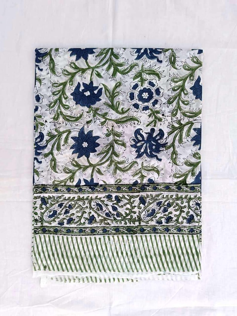 May include: A folded white textile with a repeating floral pattern in navy blue and green. The design features stylized flowers and leaves. A border with paisley and striped patterns is visible.