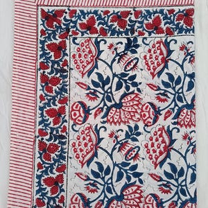 May include: White textile with a repeating floral pattern in red and blue. The design features large stylized flowers and leaves. A border of red and white stripes frames the edge, creating a cohesive look. The fabric is likely cotton or a similar material.