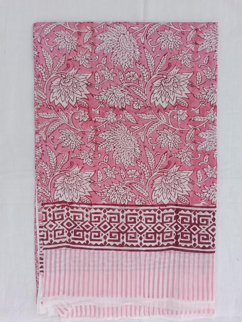 May include: Pink floral fabric featuring a white floral design. The fabric has a border with geometric shapes in a darker pink and a striped pattern. Suitable for home decor or clothing, the fabric is likely cotton or linen.