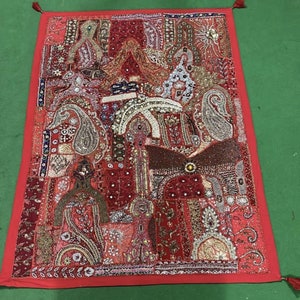 Puede incluir: Un tapiz de pared rojo con bordados e incrustaciones intrincados. El tapiz presenta un diseño de patchwork con varios patrones y texturas, incluyendo motivos paisley, florales y geométricos. El tapiz está adornado con cuentas, lentejuelas y otros elementos decorativos.