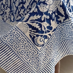 May include: A blue and white patterned tablecloth with a floral design. The tablecloth has a border with geometric shapes and stripes. The fabric is draped over a wooden table, showcasing the intricate details of the textile.