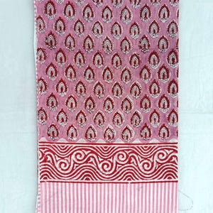 May include: Pink fabric with a repeating pattern of red and white teardrop shapes. The bottom border has a red wavy design above a striped section. The fabric is likely cotton or linen, suitable for textile projects.