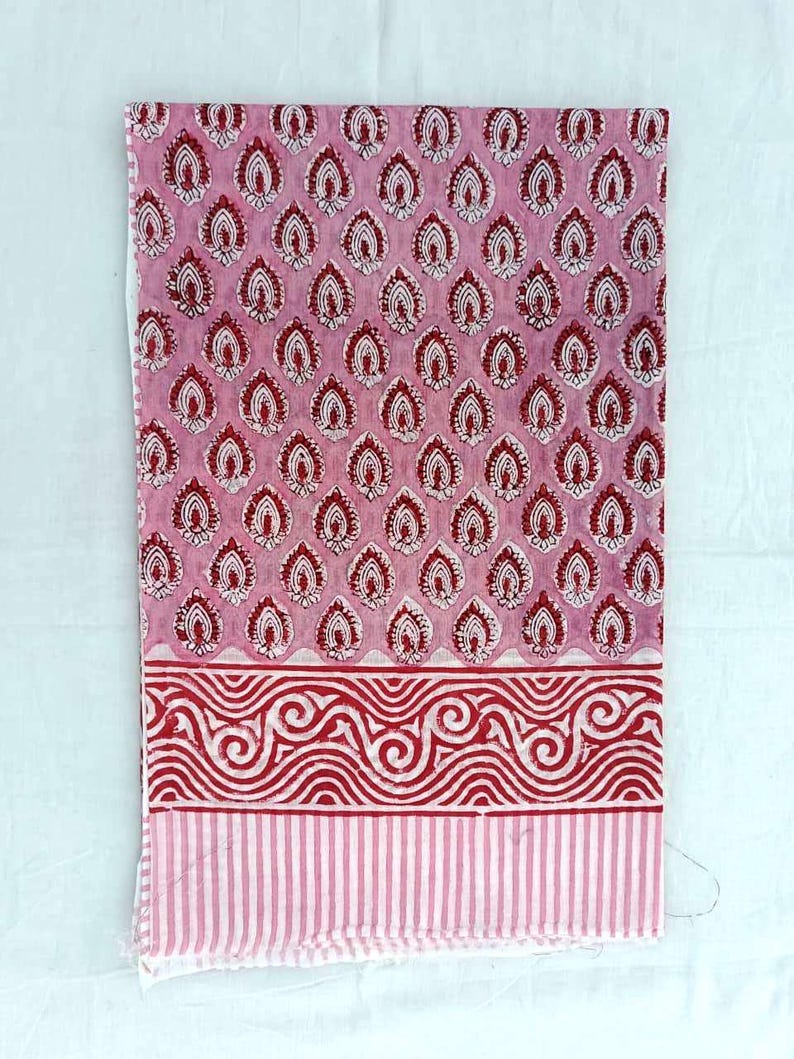 May include: Pink fabric with a repeating pattern of red and white teardrop shapes. The bottom border has a red wavy design above a striped section. The fabric is likely cotton or linen, suitable for textile projects.
