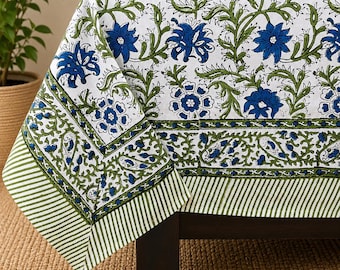 Può includere: Una tovaglia bianca con un motivo floreale ripetuto in blu e verde. Il design presenta grandi fiori blu, viticci verdi e un bordo a righe verdi e bianche, drappeggiato sull'angolo di un tavolo in legno scuro.
