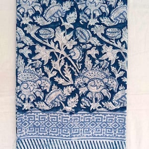 May include: A folded textile with a repeating floral pattern in white on a dark blue backdrop. The design features stylized flowers and leaves. A border with geometric shapes and stripes is at the bottom.