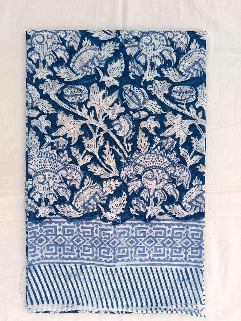 May include: A folded textile with a repeating floral pattern in white on a dark blue backdrop. The design features stylized flowers and leaves. A border with geometric shapes and stripes is at the bottom.